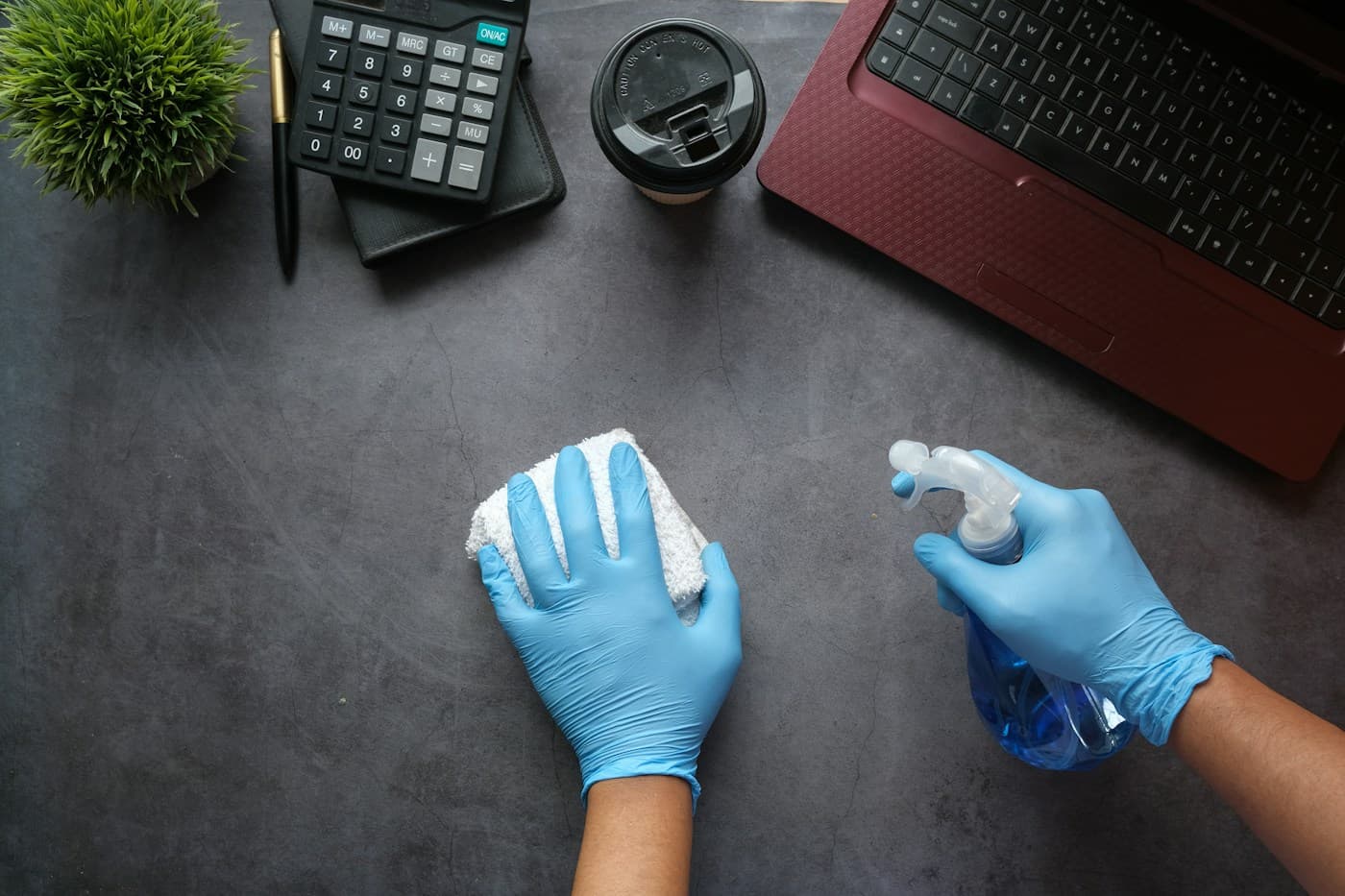 Blue gloved hands spray and wipe a desk with a cloth